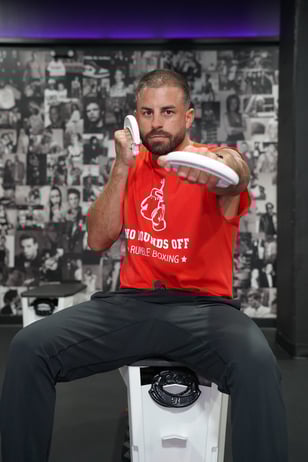 Male trainee wearing a red tshirt displaying Rumble Boxing motto saying "no rounds off" while throwing a shadow punch forward while holding weights in hand